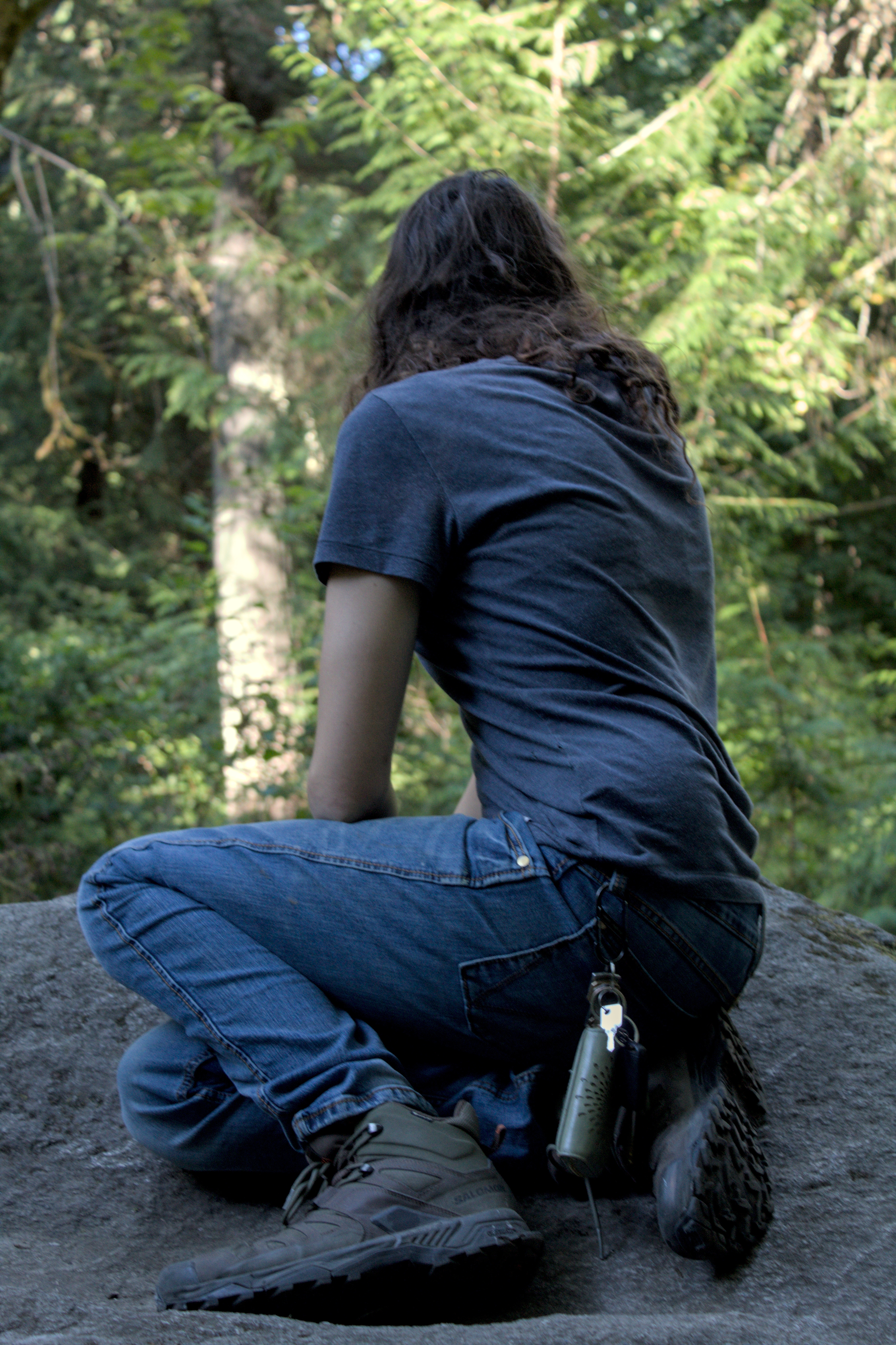 white woman in a tshirt, jeans, and hiking boots squats on a rock, facing away from the camera and towards the trees in the background.